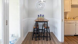 Uma sala de jantar com uma mesa de madeira e duas cadeiras, um relógio na parede e uma cozinha visível através de uma porta aberta.