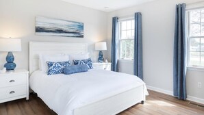 Quarto com cama branca, duas mesas de cabeceira, janela com cortinas e um quadro na parede.