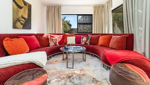 A modern living room with a curved red sofa, a round coffee table, and a large window with curtains. - Charming Garden Cottage-campsbay (Cape Town)