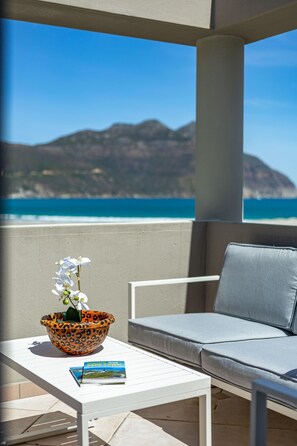 Uma varanda com uma mesa branca, um sofá, uma tigela com estampa de leopardo e uma orquídea, e um livro. Ao fundo, vista do oceano e montanhas.