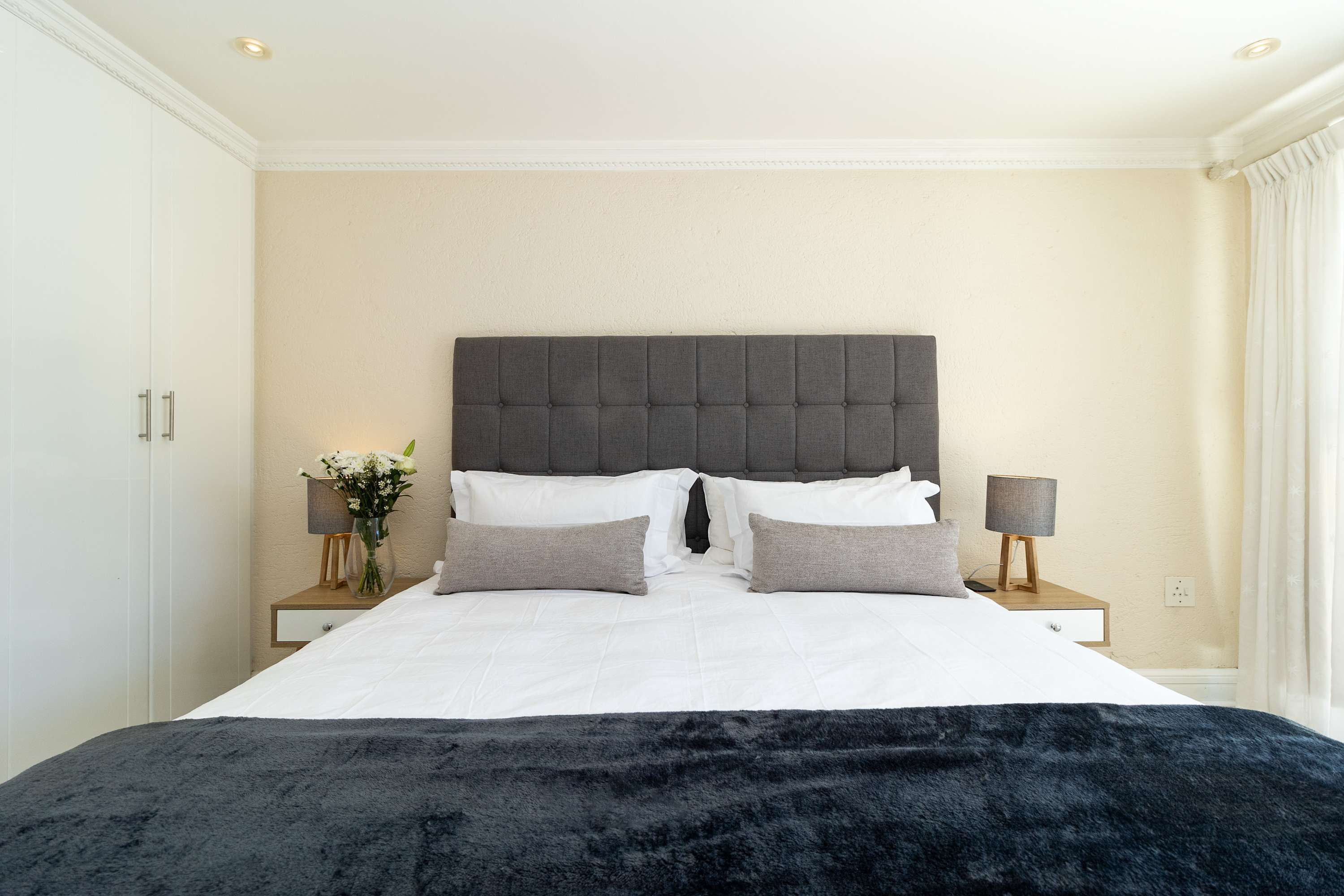 A neatly made bed with a grey headboard, white bedding, and a black throw blanket. There are two bedside tables with lamps and a vase of flowers.