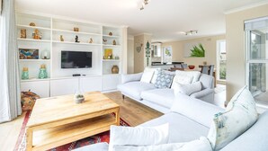 A modern living room with a white sofa, a wooden coffee table, and a TV mounted on a white cabinet. - 3 Bedroom Family Home - Houtbay (Cape Town)