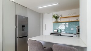 A modern kitchen with a stainless steel refrigerator, a white countertop, and two grey chairs.