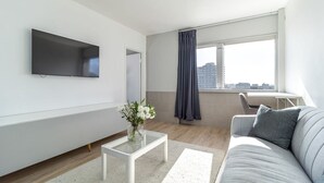 A modern living room with a flat-screen TV, a glass coffee table with a vase of flowers, a grey sofa, and a large window with a view of buildings.