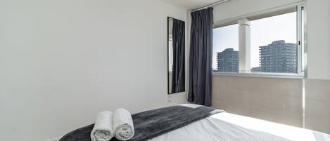 A modern hotel room with a large bed, a window with curtains, and a view of city buildings.
