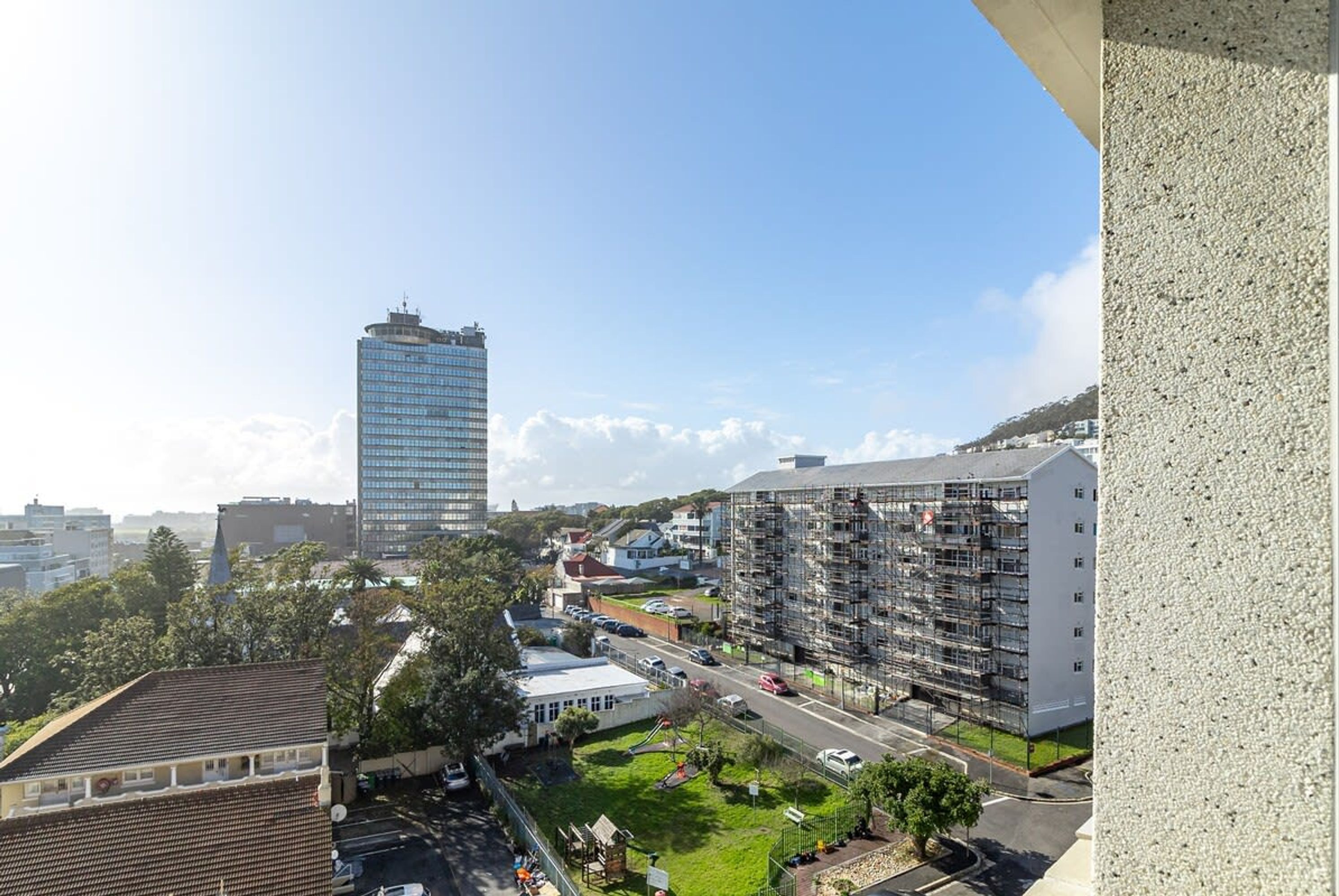 Un edificio de gran altura con fachada de vidrio moderna, un edificio residencial en construcción con andamios, y una calle con automóviles y vegetación.