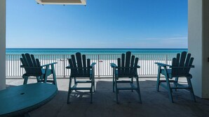 A balcony with three Adirondack chairs and a table, overlooking a beach with a clear blue sky and ocean.
