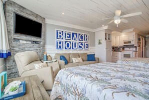 A bedroom with a large bed, a television mounted on a brick wall, and a kitchen area visible through an open door. - Seaside Villa | Ocean View, Pool | Activity Experiences (Panama City Beach)