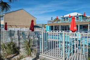A pool area with a fenced deck, red umbrellas, and a building with a blue roof in the background. - The Turtle Nest | Pool, Beach Access | Activity Experiences (Panama City Beach)