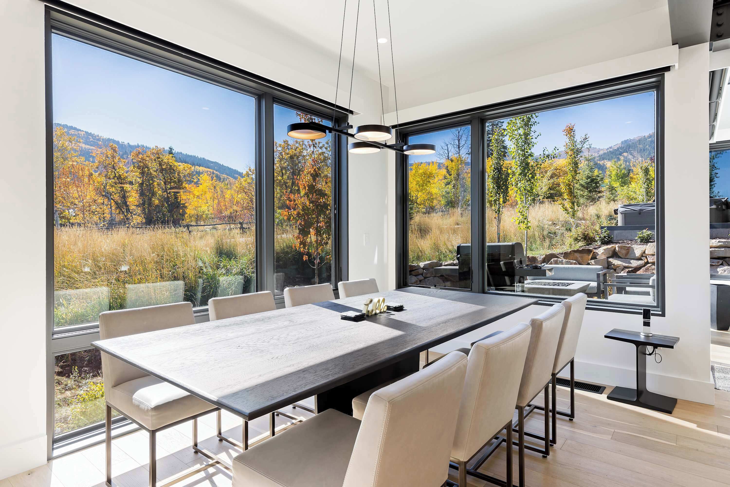 Un comedor moderno con una mesa grande, varias sillas y ventanas amplias que ofrecen vistas a un paisaje boscoso.