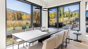 Een moderne eethoek met een grote tafel, meerdere stoelen en grote ramen met uitzicht op een bebost landschap.