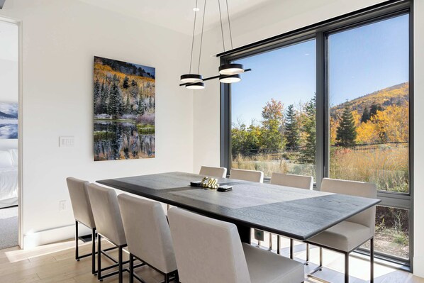 Un comedor moderno con una mesa grande, sillas blancas y un cuadro de un paisaje forestal.