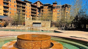 A pool with a central fountain, surrounded by a stone deck and a wooden building in the background. - Serenity King | Balcony (Park City)