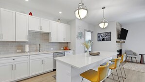 Una cocina moderna con armarios blancos, una isla grande con taburetes y luminarias colgantes, además de un televisor de pantalla plana en la pared.