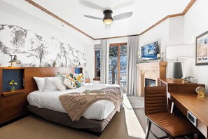 A bedroom with a bed, a desk, a chair, a ceiling fan, a fireplace, and a large window.