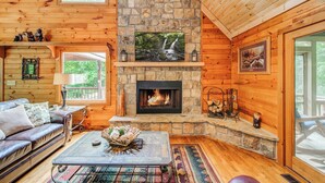 Un interior de cabaña acogedor, con una chimenea de piedra, paredes de madera, un sofá cómodo y un televisor sobre la chimenea.
