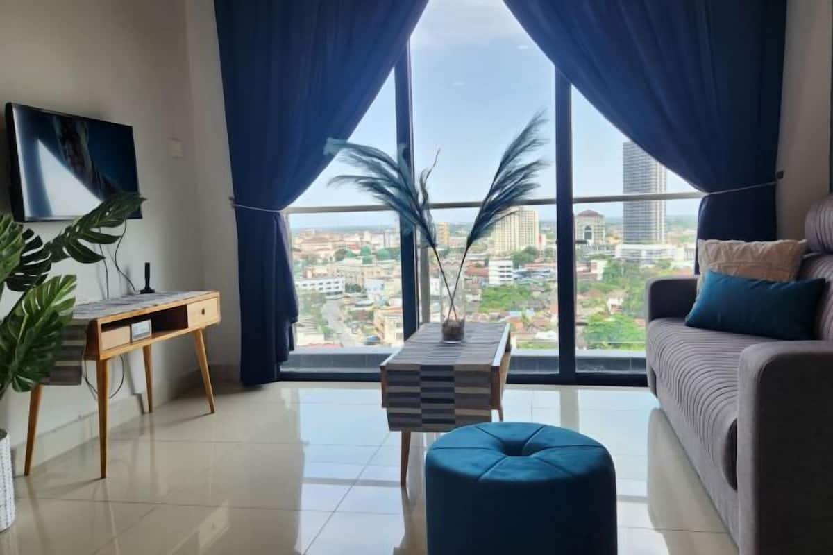 Una sala de estar moderna con un sofá, un escritorio y vistas a un paisaje urbano a través de amplios ventanales.