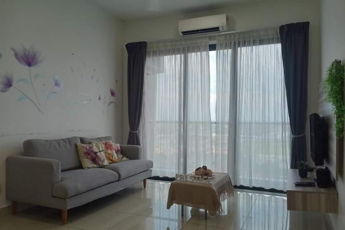 Una sala de estar con un sofá, una mesa de centro, un televisor montado en la pared y una ventana con cortinas translúcidas.