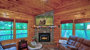 A cozy cabin interior with a stone fireplace, wooden walls, and large windows offering forest views. - Pet-Friendly escape in Ellijay! Private Hot Tub! (Ellijay)