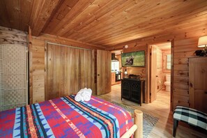 A wooden cabin room with a bed, a television, a desk, and a bathroom visible through an open door. - The Guest Cabin - Historic Home in Big Canoe (Big Canoe)