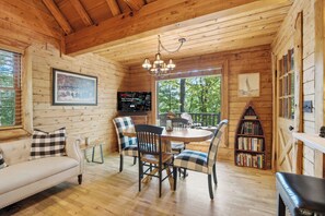 A cozy cabin interior with a wooden ceiling, a dining table with chairs, a sofa, a bookshelf, and a television mounted on the wall. - The Guest Cabin - Historic Home in Big Canoe (Big Canoe)