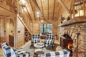 A log cabin interior with a stone fireplace, checkered furniture, and a wooden staircase.