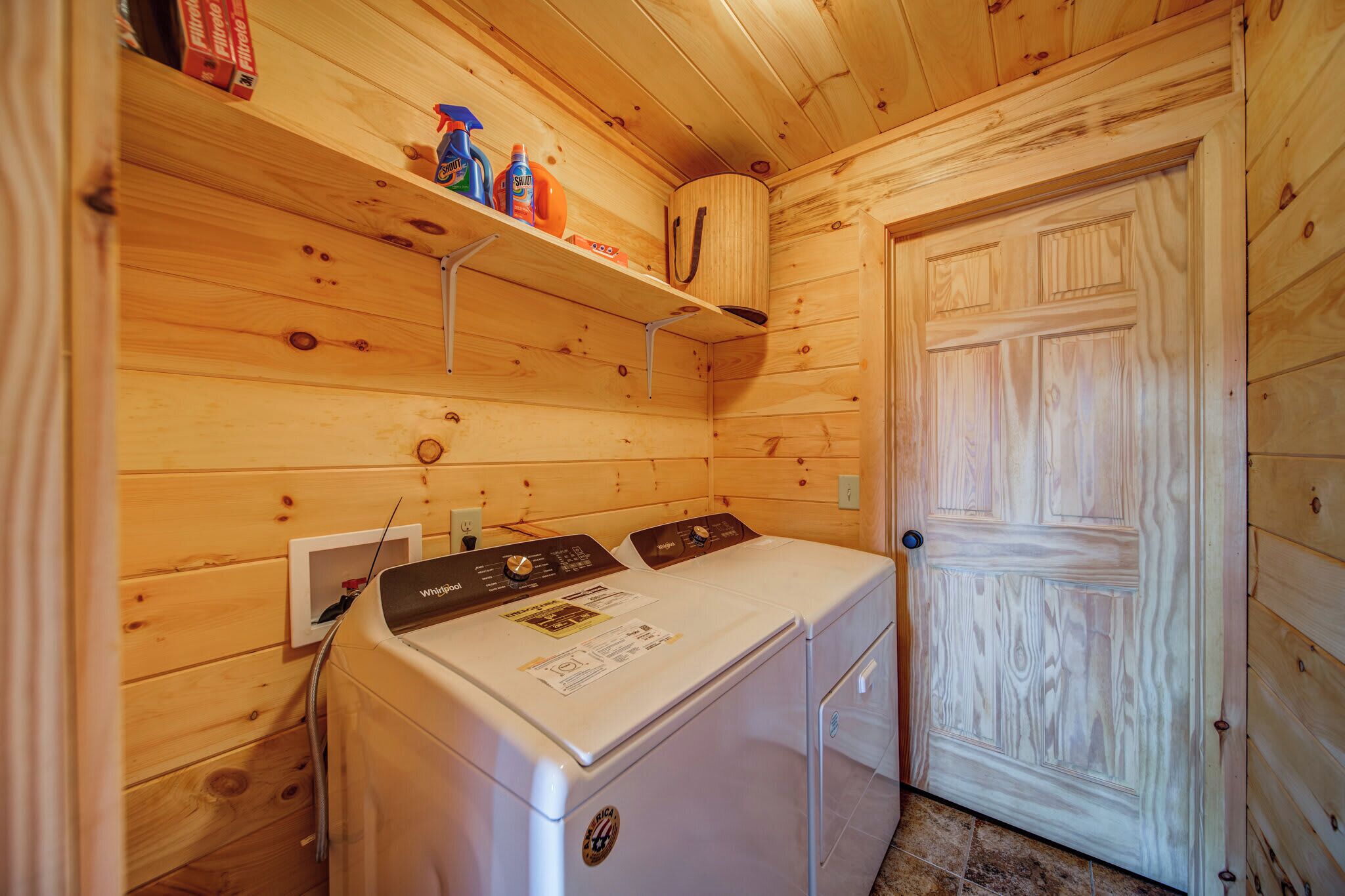 A compact laundry room with a front-loading washer and dryer, wooden walls, and a shelf with cleaning supplies.