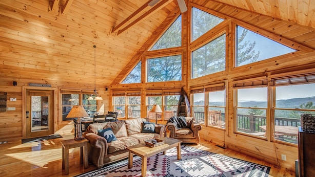 Een ruime houten chalet met grote ramen, een gezellige zithoek met een bank en fauteuils, en uitzicht op het omliggende landschap.