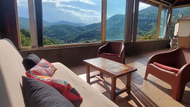 Ein Zimmer mit Holzboden, großen Fenstern mit Bergblick, einer Couch mit bunten Kissen und einem kleinen Tisch.