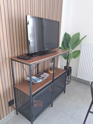En tv-apparat står på ett TV-bord i trä. Bredvid den ligger en tidning, en fjärrkontroll och en krukväxt.