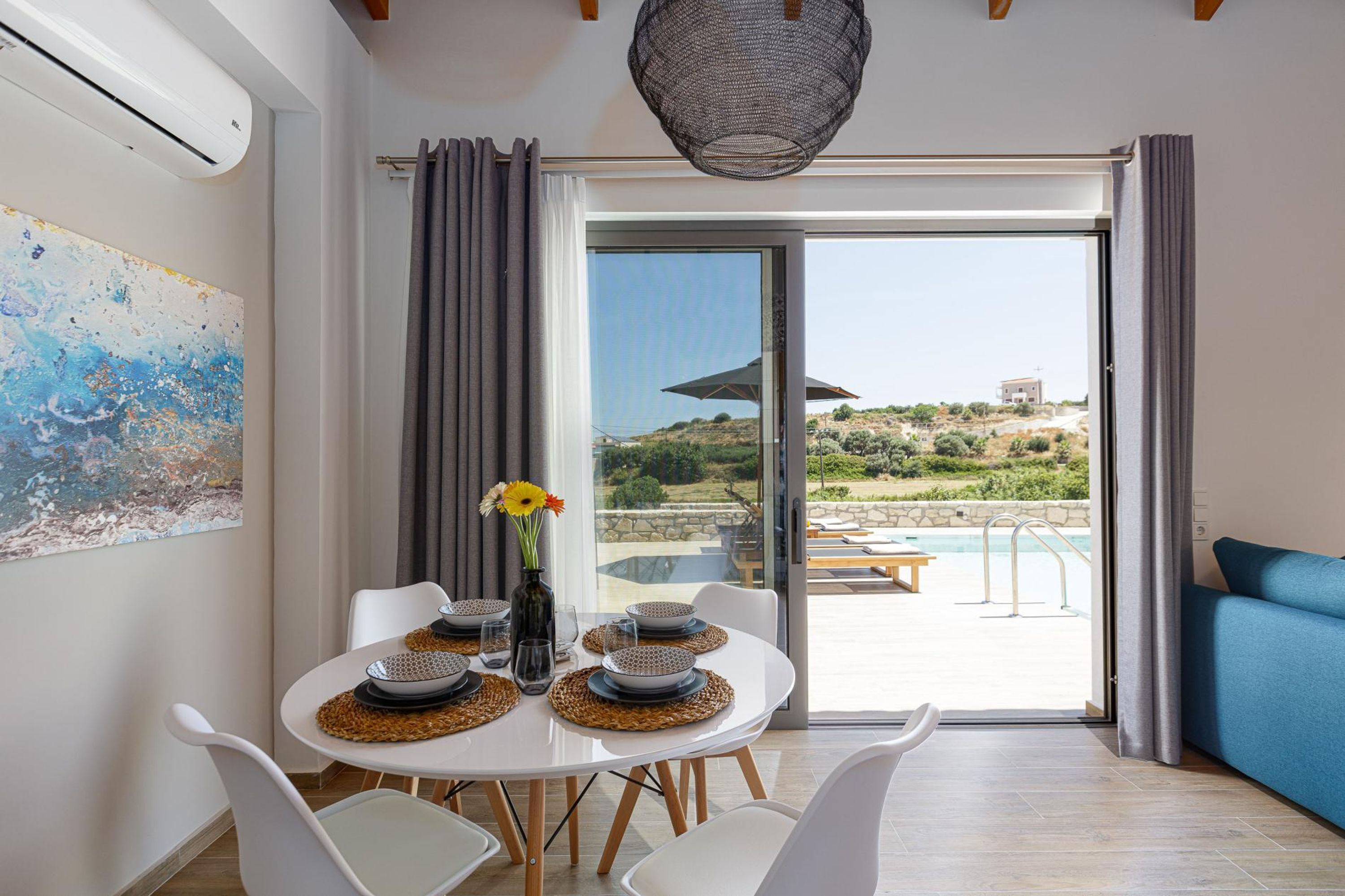 Un comedor con una mesa preparada para cuatro, un cuadro abstracto grande, y vista a una piscina y paisaje a través de una puerta de cristal.