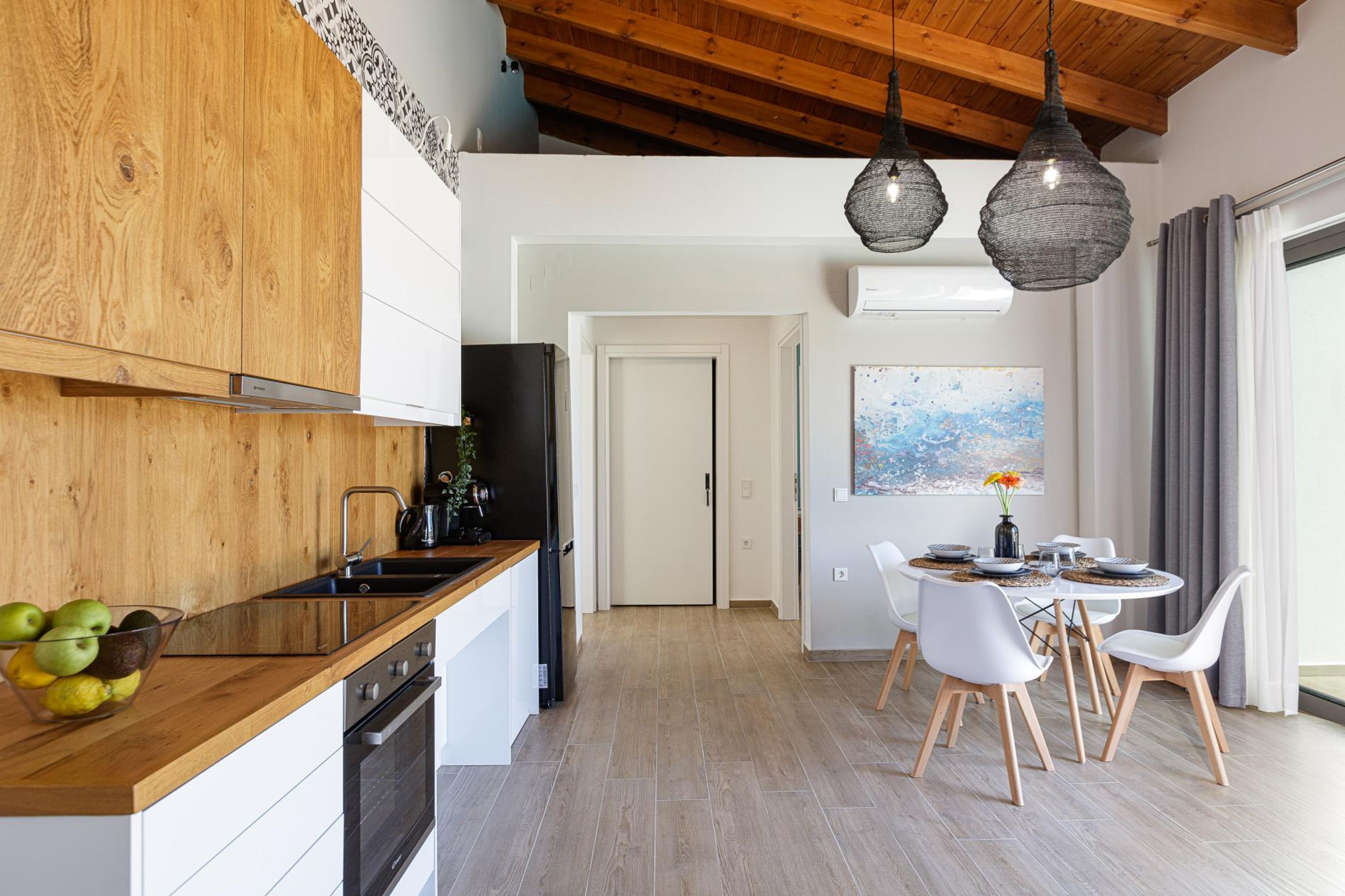 Una cucina moderna con mobili in legno, un piano cottura nero e una ciotola di frutta sul bancone. C'è una zona pranzo con sedie bianche e un tavolo, e un quadro appeso al muro.