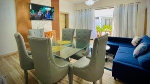 Uma sala de estar moderna com uma mesa de jantar de vidro, cadeiras estofadas cinzentas, um sofá azul e uma televisão de tela plana fixada na parede.