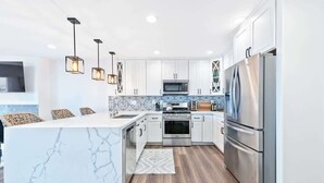 Cocina moderna con armarios blancos, una isla de mármol, electrodomésticos de acero inoxidable e iluminación colgante.