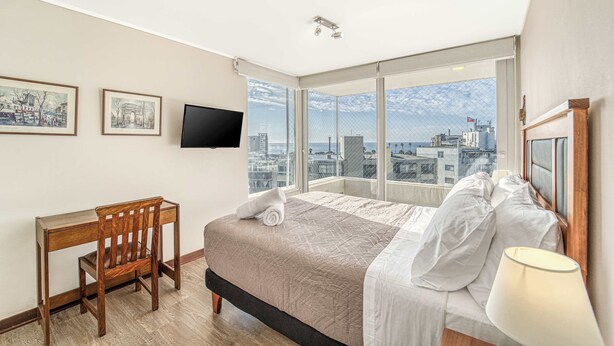 A hotel room with a bed, a desk, a chair, a TV, and a large window with a city view. - Costa Vina 3B  Balcony & View (Viña del Mar)