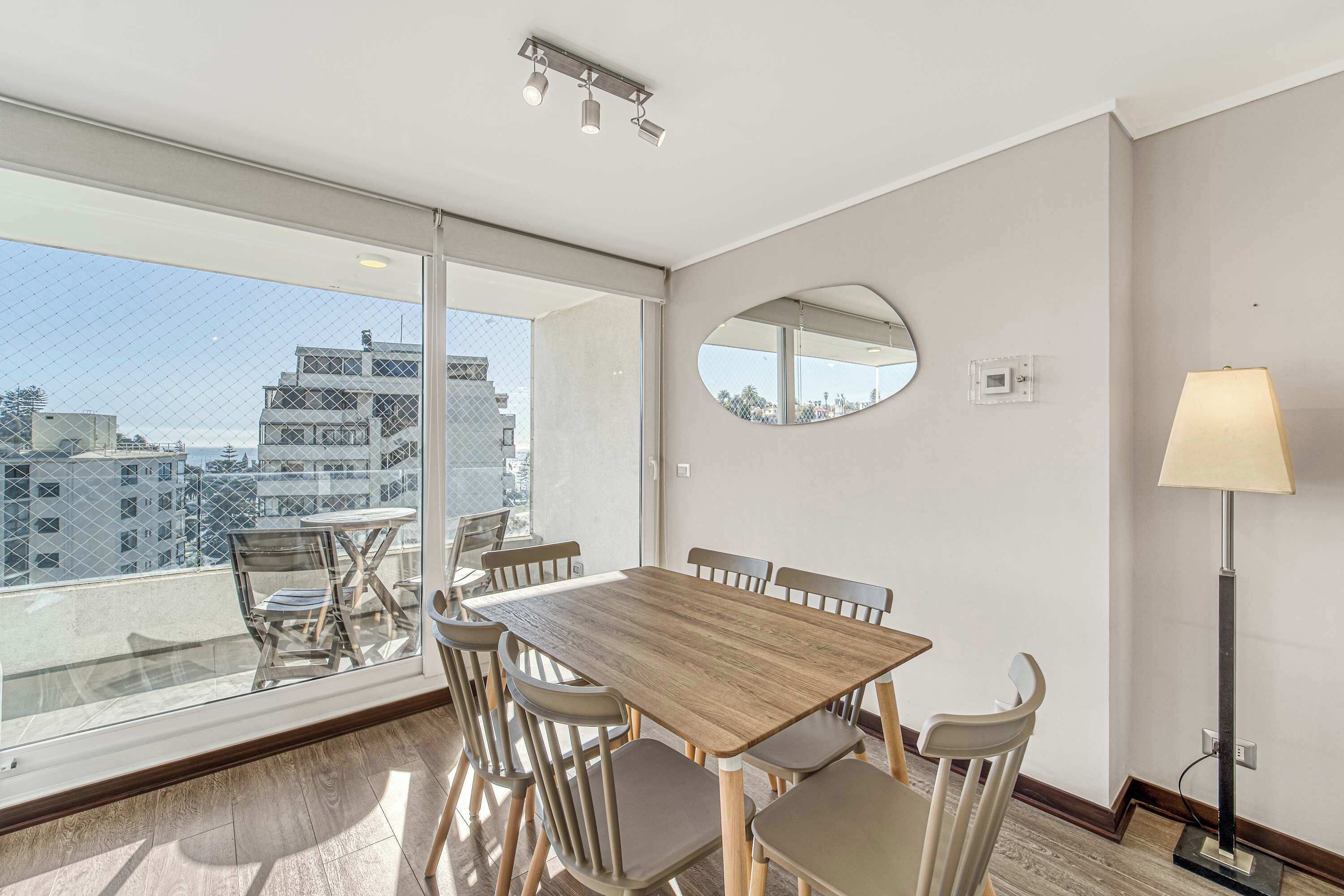 Una habitación moderna con un ventanal grande, un espejo redondo, una mesa con sillas y una lámpara de pie.