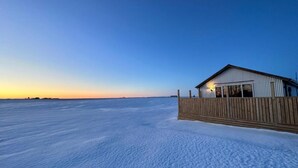 A snow-covered landscape with a wooden cabin featuring a large window and a light source inside. - Cave Cabins (Rangárþing ytra)