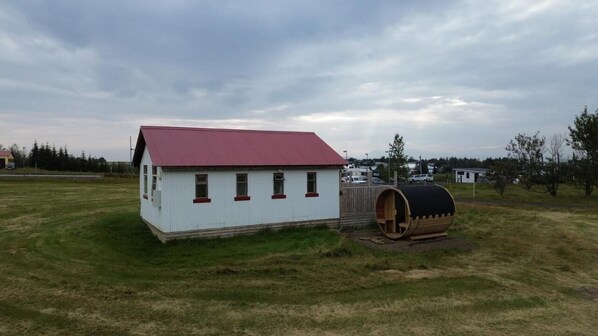A small, single-story building with a red roof and white walls, located in a grassy field with a wooden barrel nearby. - Cave Cabins (Rangárþing ytra)