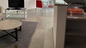 Un soggiorno moderno con una TV a schermo piatto, un tavolino in vetro e una poltrona rossa. È presente anche un angolo cottura con un fornello a gas e un tavolo da pranzo con sedie rosse.
