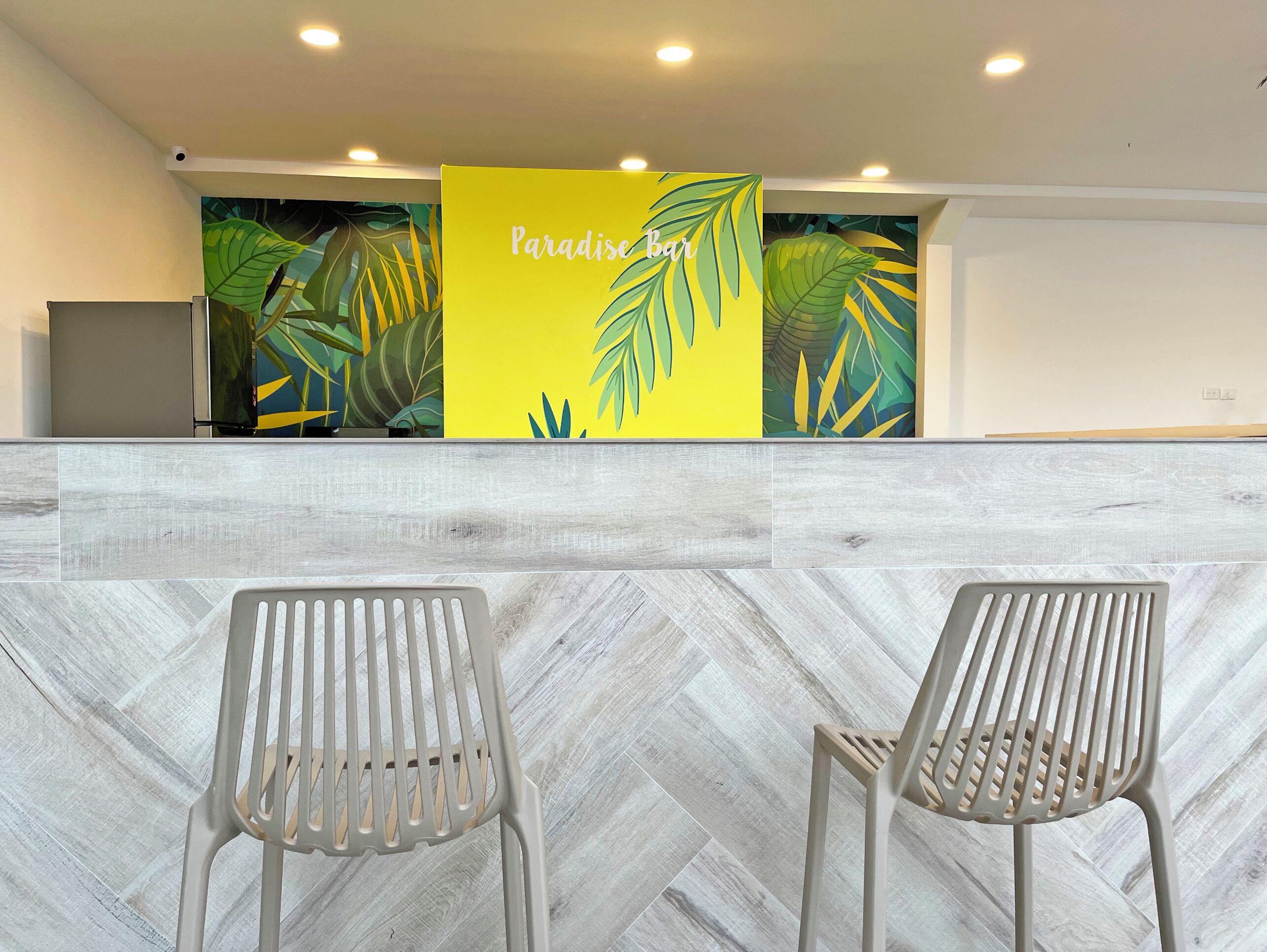 Una zona de recepción con piso de madera en patrón de espiga, dos sillas de plástico gris y una pared amarilla con motivos de hojas tropicales y la palabra 'Paradise' escrita.