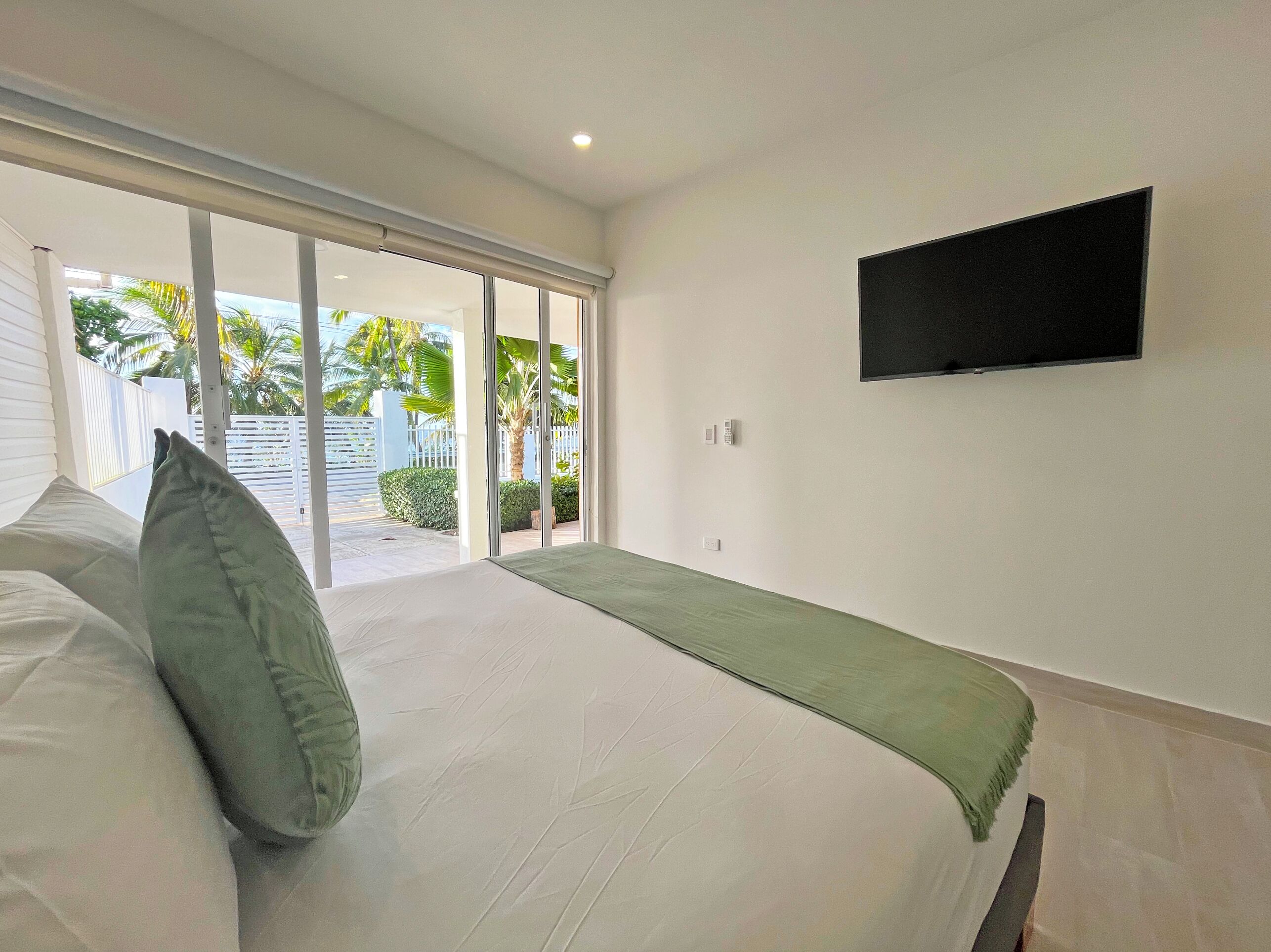 A bedroom with a bed, a TV mounted on the wall, and a sliding glass door leading to a balcony with a view of palm trees.
