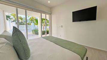 A bedroom with a bed, a TV mounted on the wall, and a sliding glass door leading to a balcony with a view of palm trees.