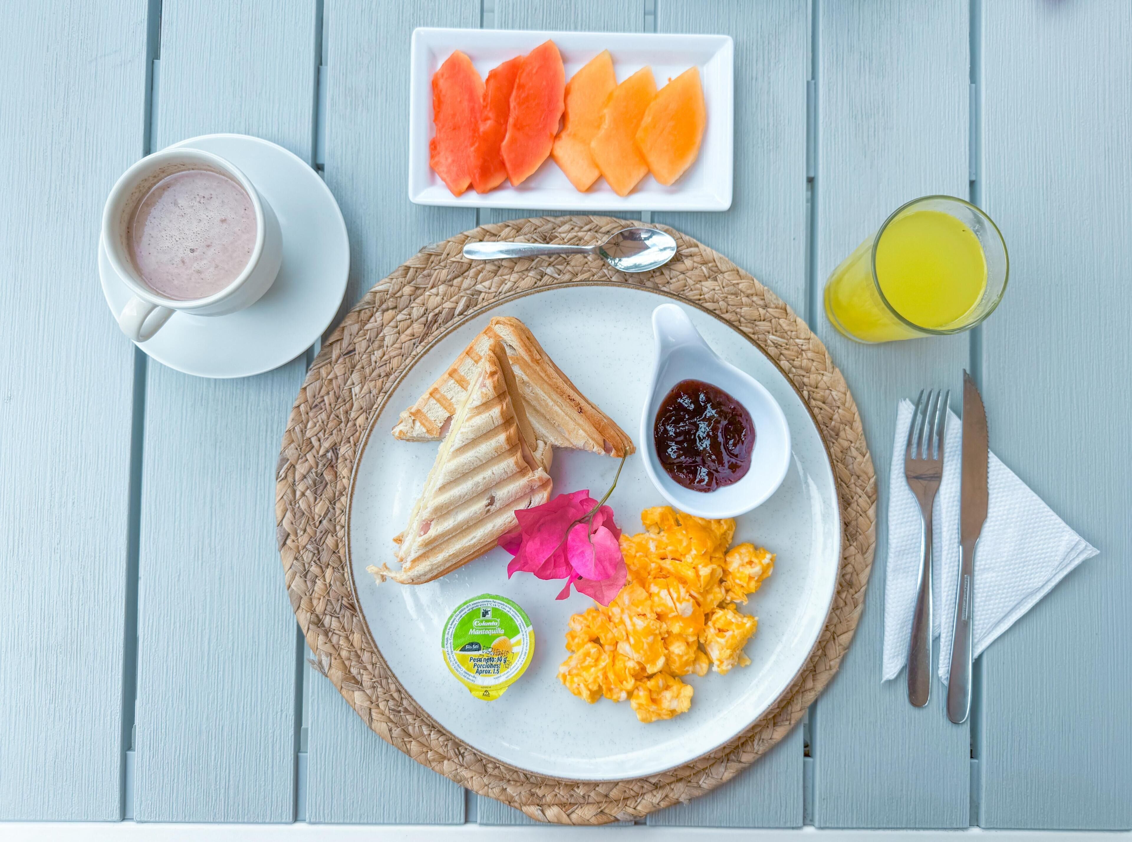 A breakfast plate with toast, scrambled eggs, a small bowl of jam, and a slice of watermelon.