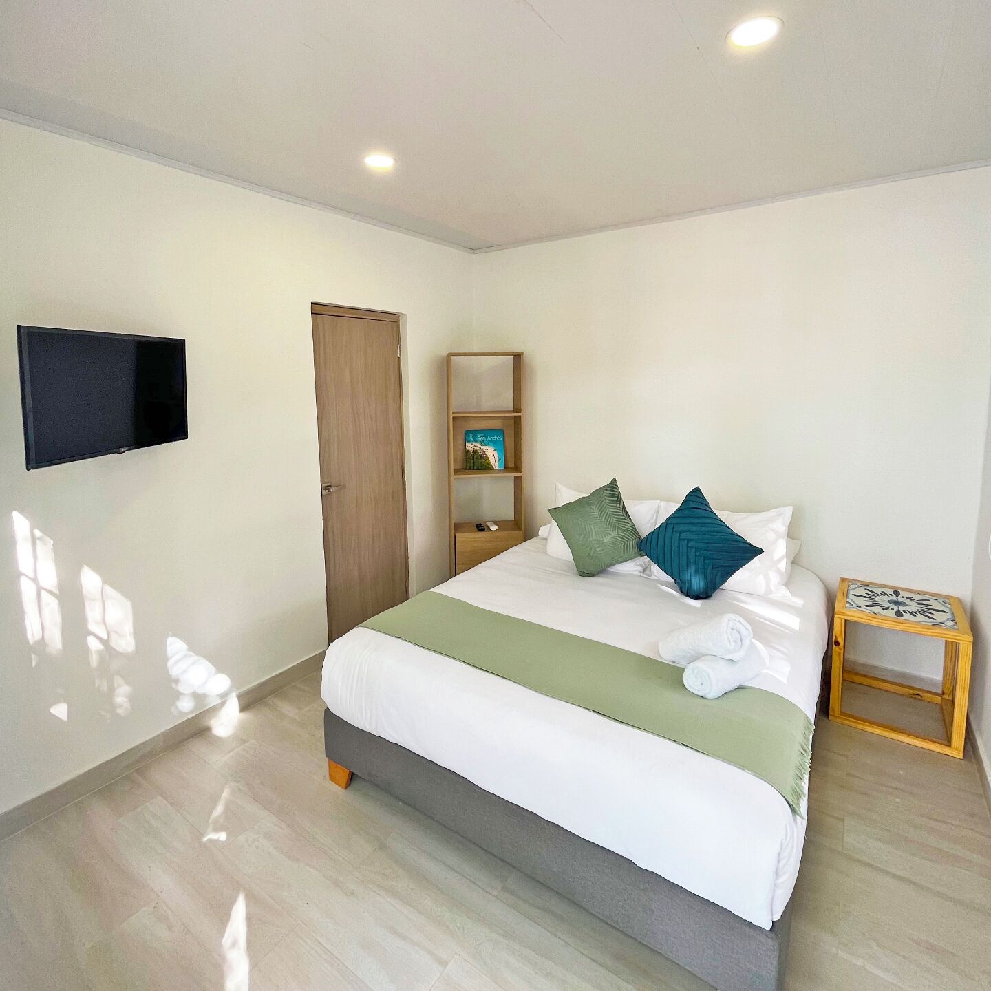 A hotel room with a bed, a TV mounted on the wall, a wooden wardrobe, a bedside table with a decorative item, and a small shelf with books.