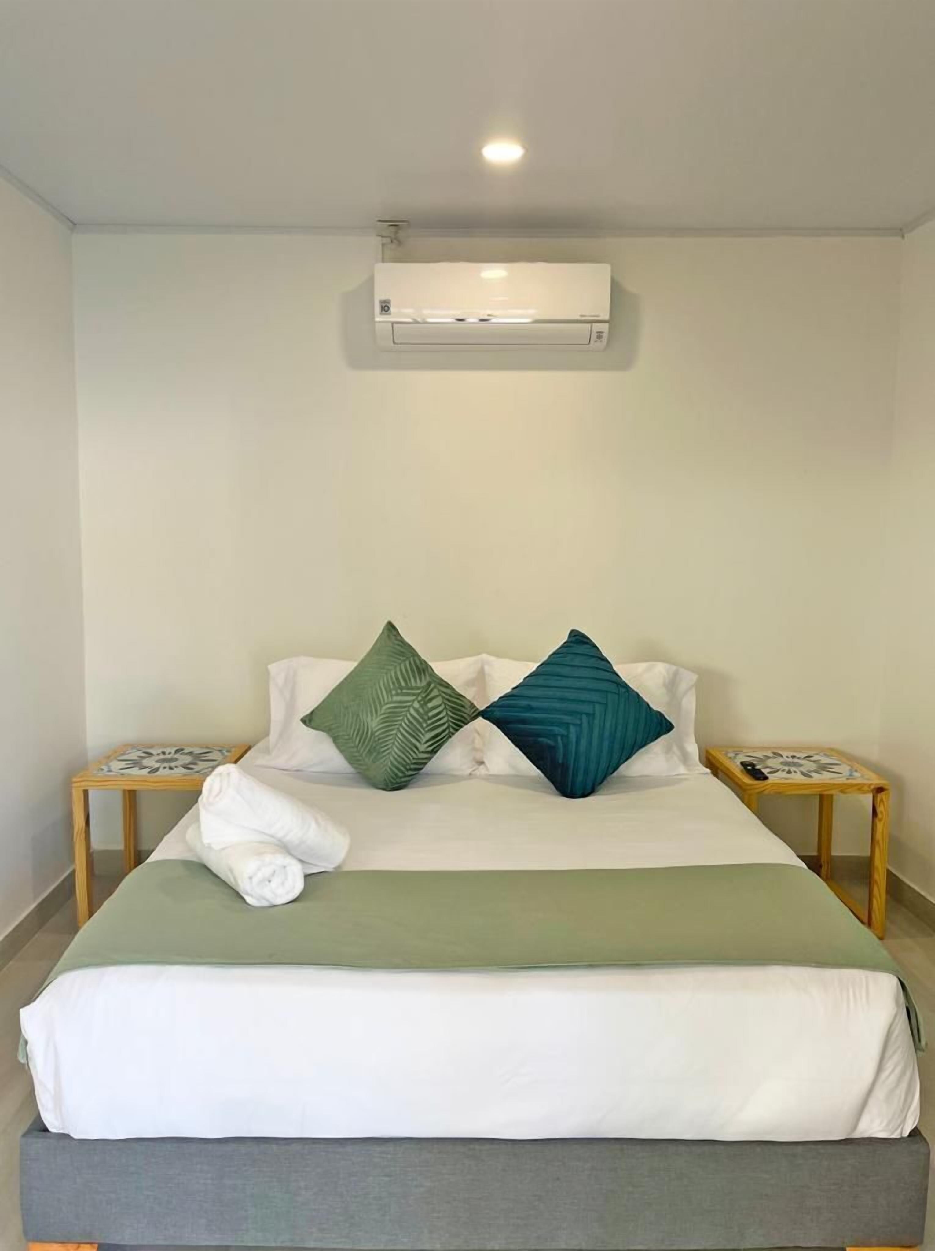 Cama bem arrumada com travesseiro verde e azul-turquesa, manta branca e toalha ao lado. Pequeno criado-mudo de madeira com luminária em cada lado da cama.