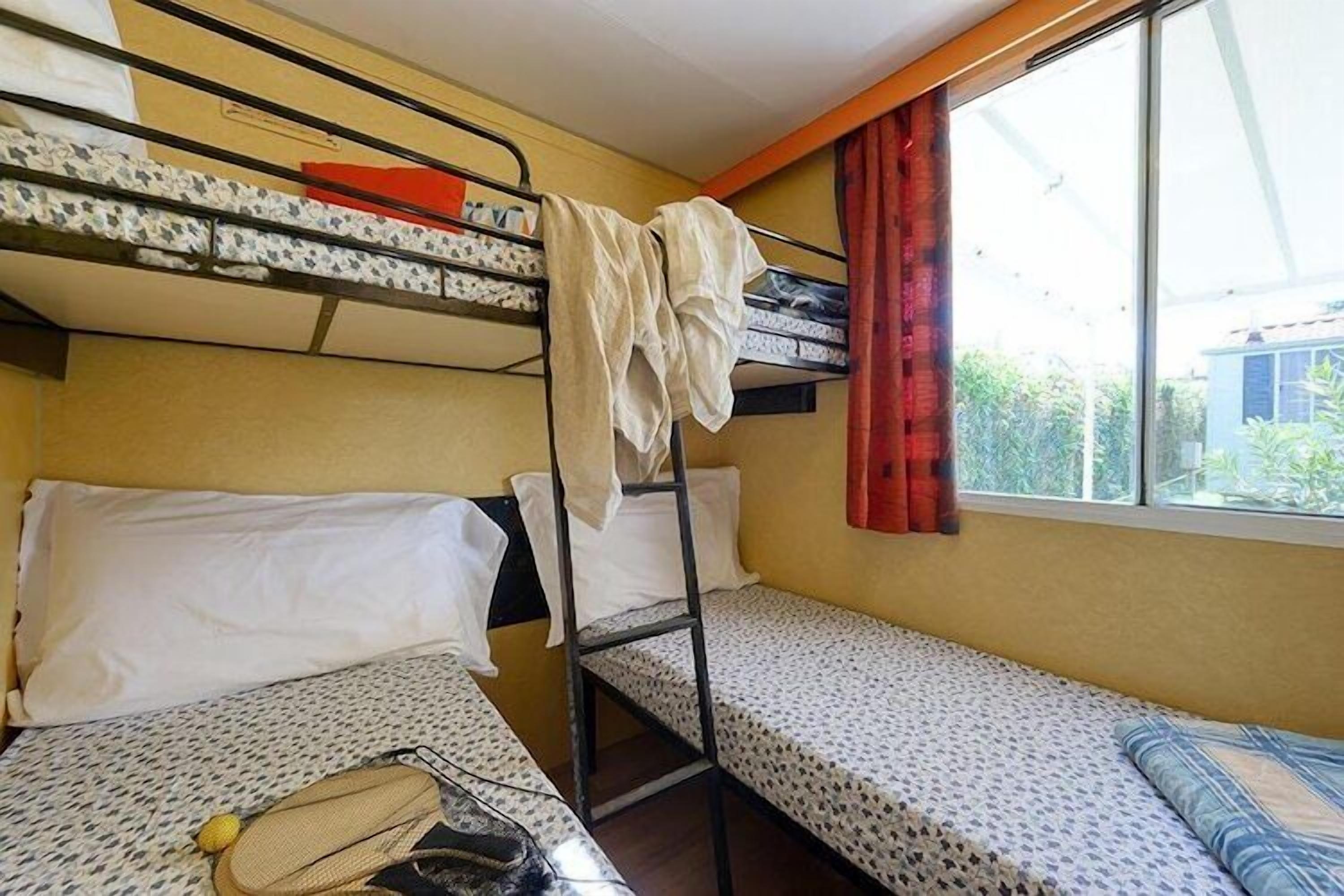 A dormitory room with bunk beds, a window with curtains, and a bed with a patterned bedspread.