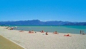 A beach with people sunbathing and swimming, clear blue water, and mountains in the background.