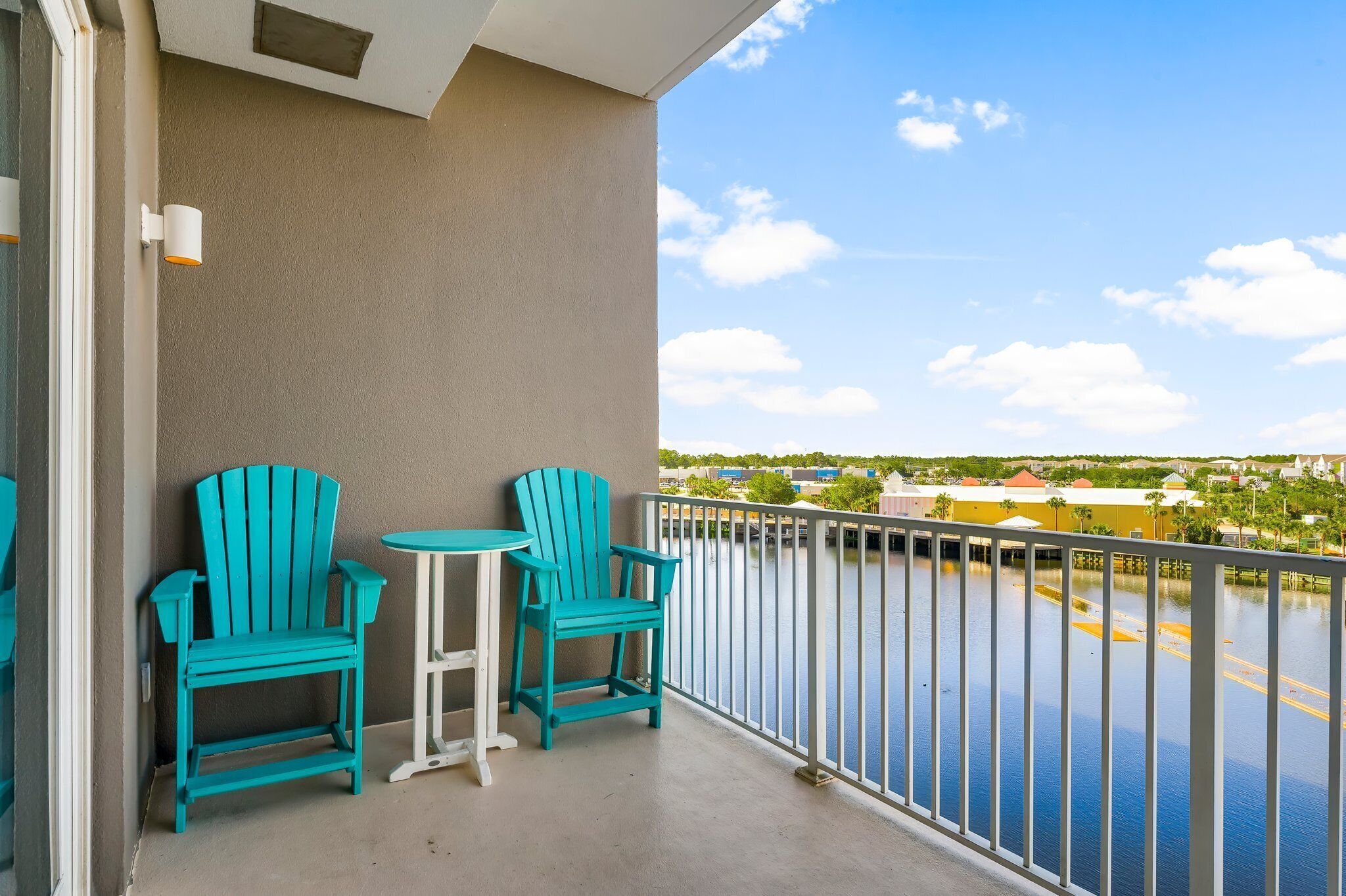 A balcony with turquoise chairs and a small table, overlooking a body of water and distant buildings.