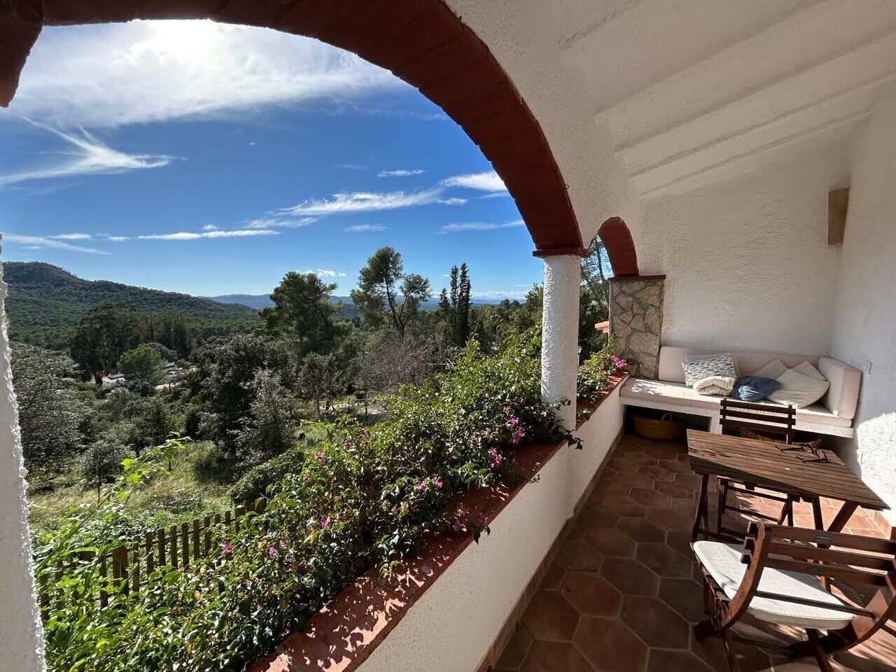 Un balcon doté d’un sol en pierre, d’une table et de chaises en bois, ainsi que d’un banc rembourré offrant une vue sur un paysage luxuriant.