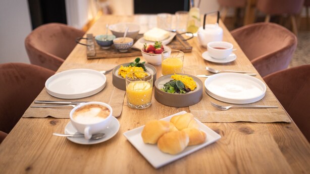 Une table en bois dressée pour le petit-déjeuner, avec des assiettes, des bols et des verres de jus d’orange.
